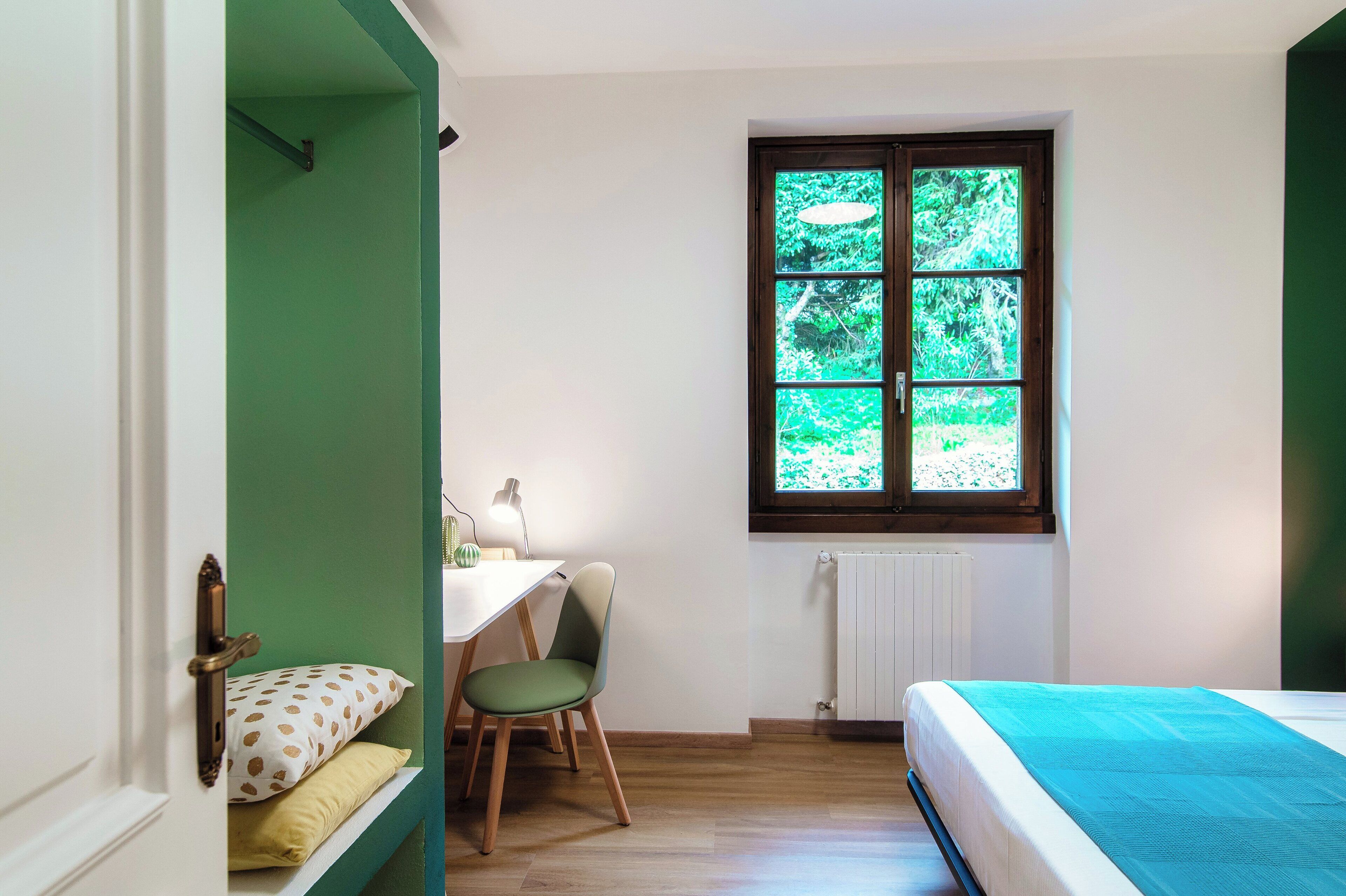 Una stanza con un letto, una scrivania con una sedia, una finestra vista sul verde e un termosifone.