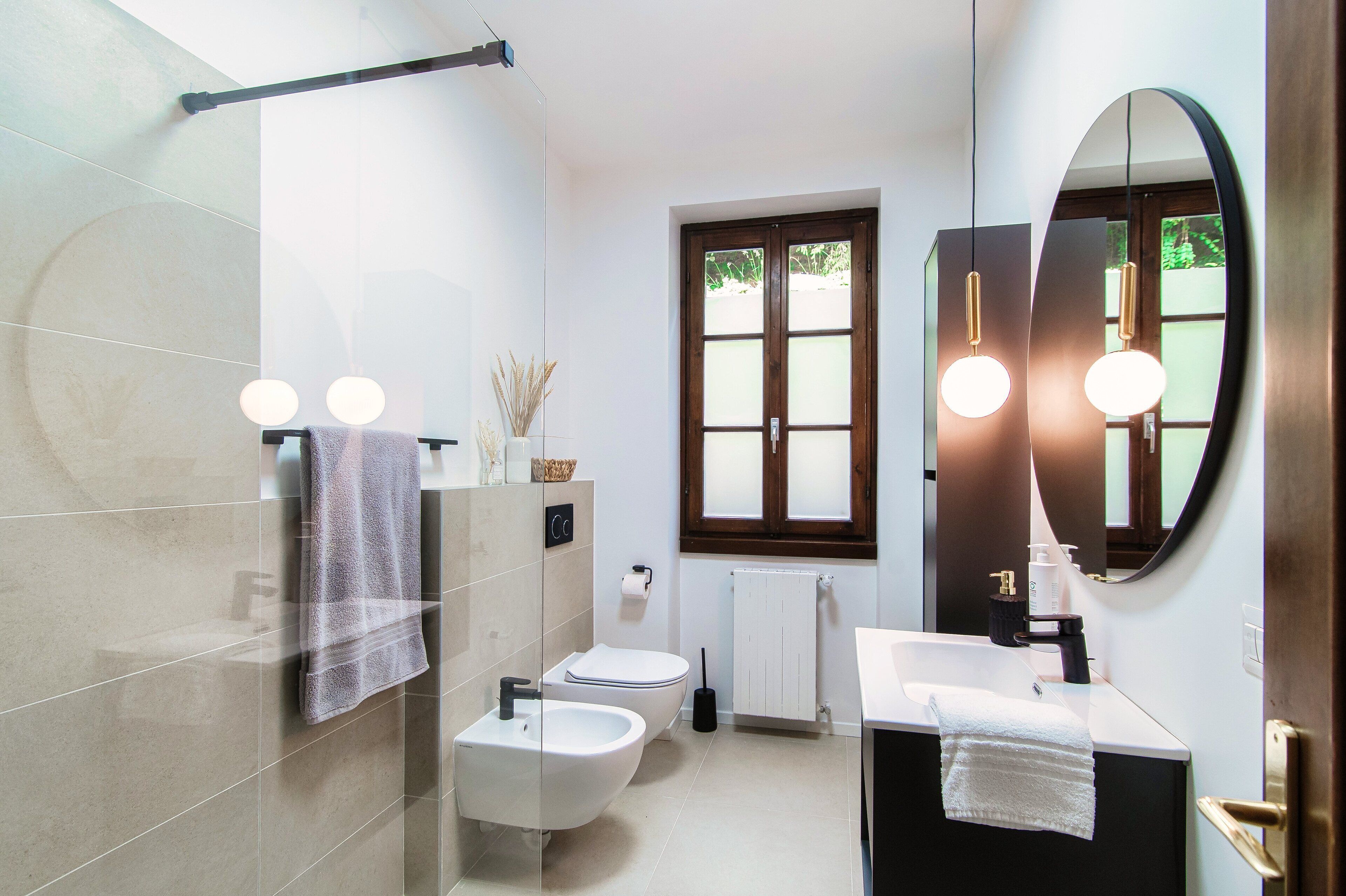 A modern bathroom with a bidet, toilet, and sink. There is a window with frosted glass, a towel rack, and a mirror.