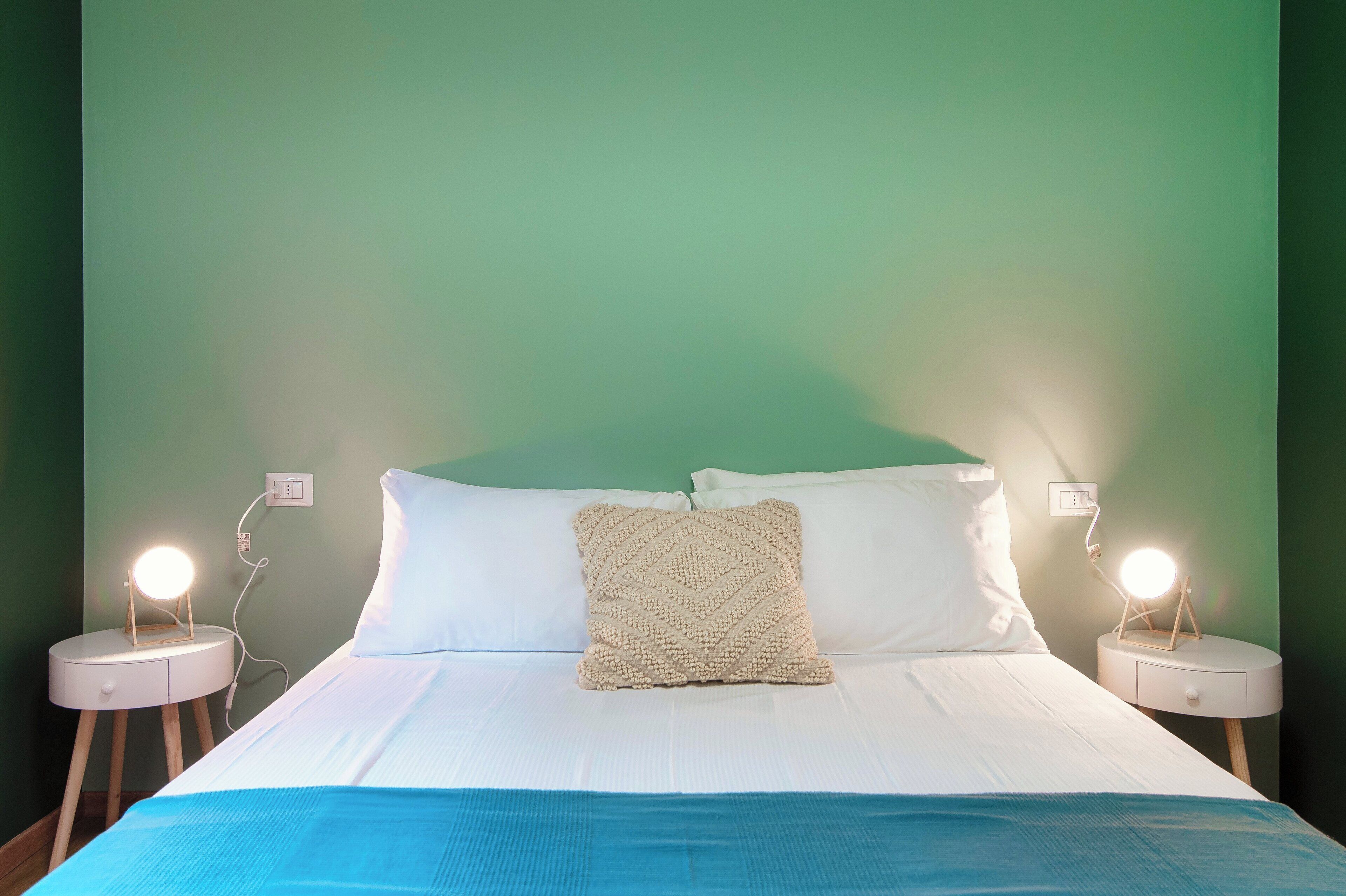 Ein Bett mit weißer Bettwäsche, einer blauen Tagesdecke und einem beige gefarbten, strukturierten Kopfkissen. Zwei Nachttische mit beleuchteten Lampen stehen daneben.