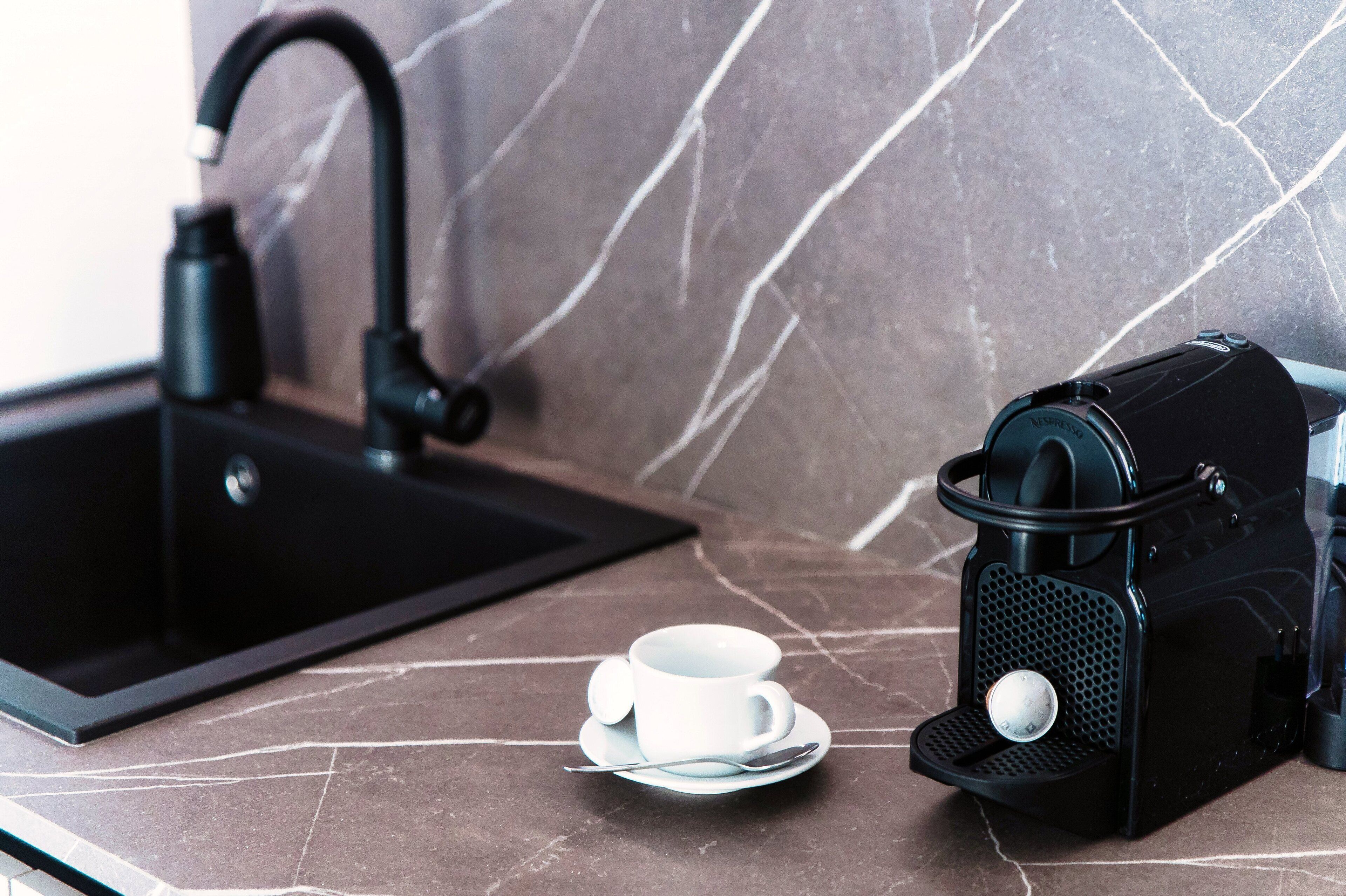 A kitchen with a black sink, a coffee machine, and a white cup on a marble countertop.