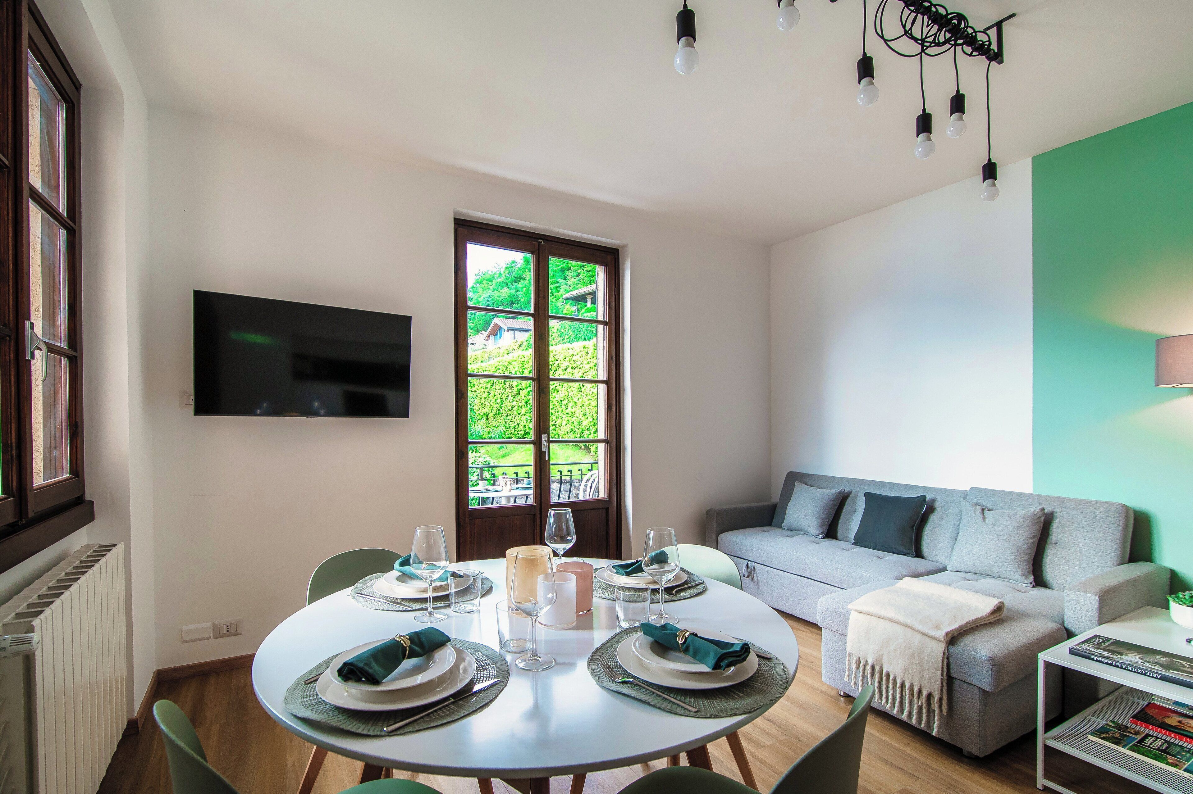 A dining area with a round table set for four, a grey sofa, and a television mounted on the wall.