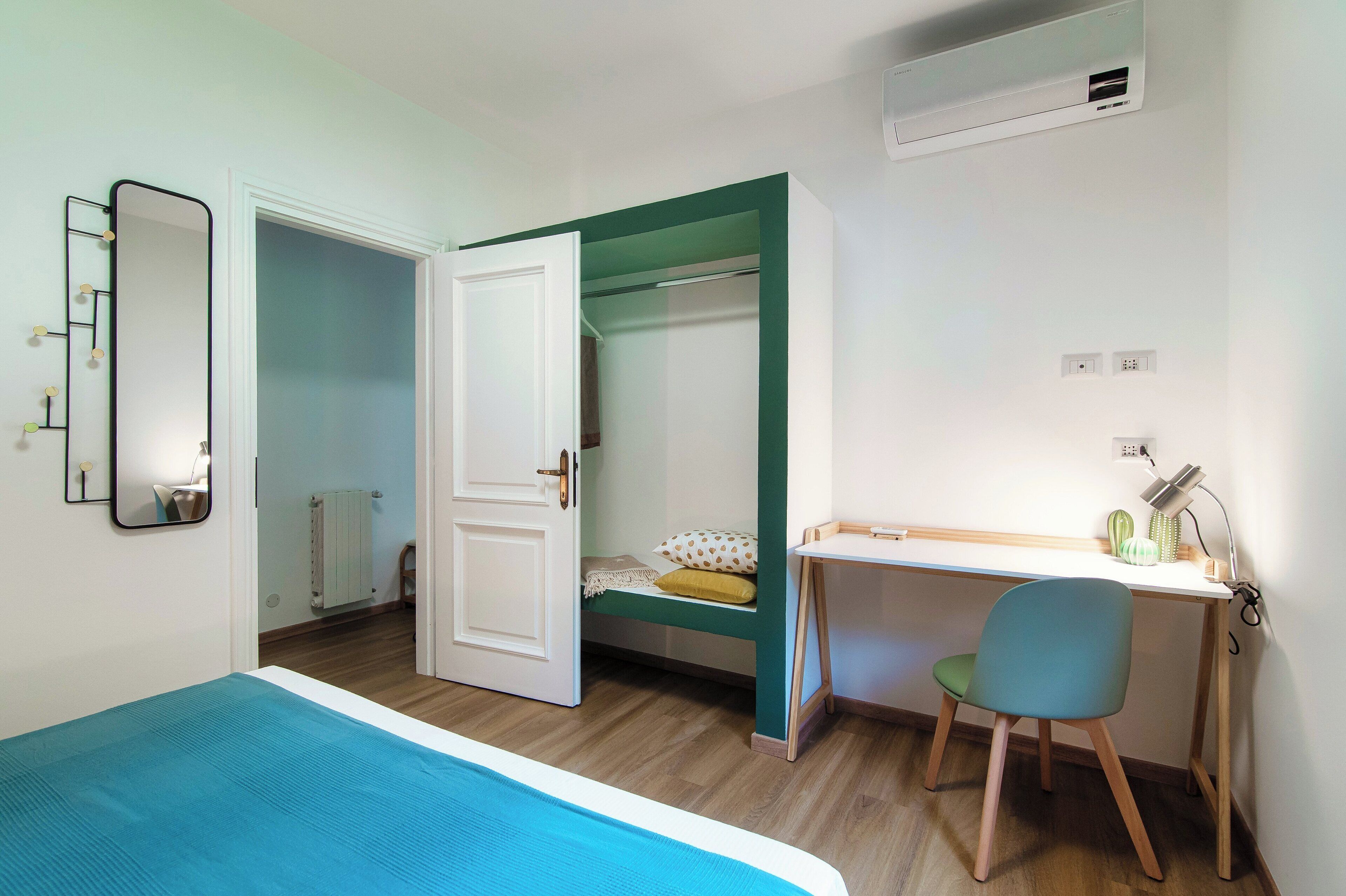 A bedroom with a bed, a desk, and a chair. There is a mirror on the wall, a radiator, and a door leading to another room.