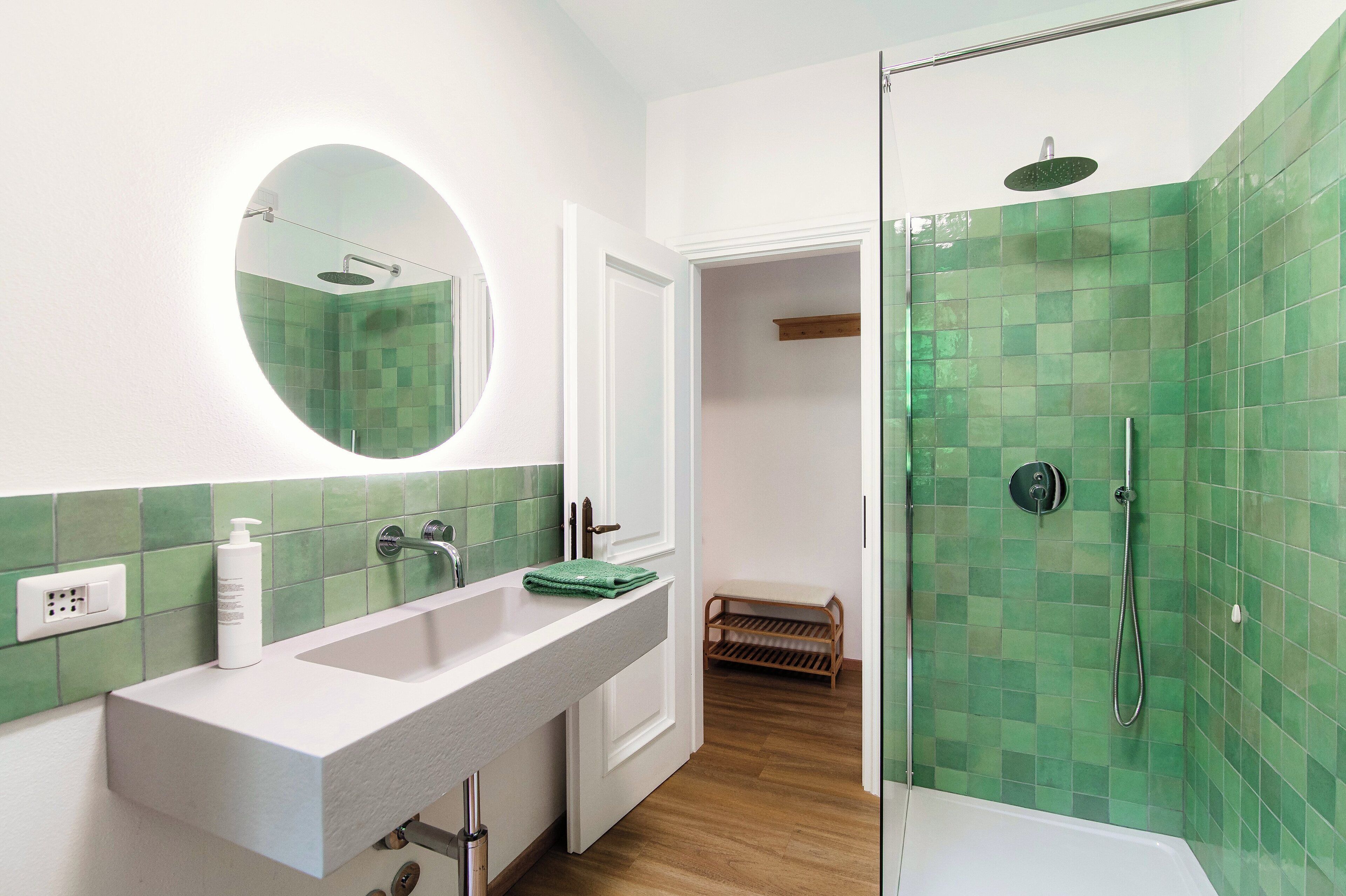 Casa de banho com lavatório branco, chuveiro com azulejos verdes e espelho sobre o lavatório.