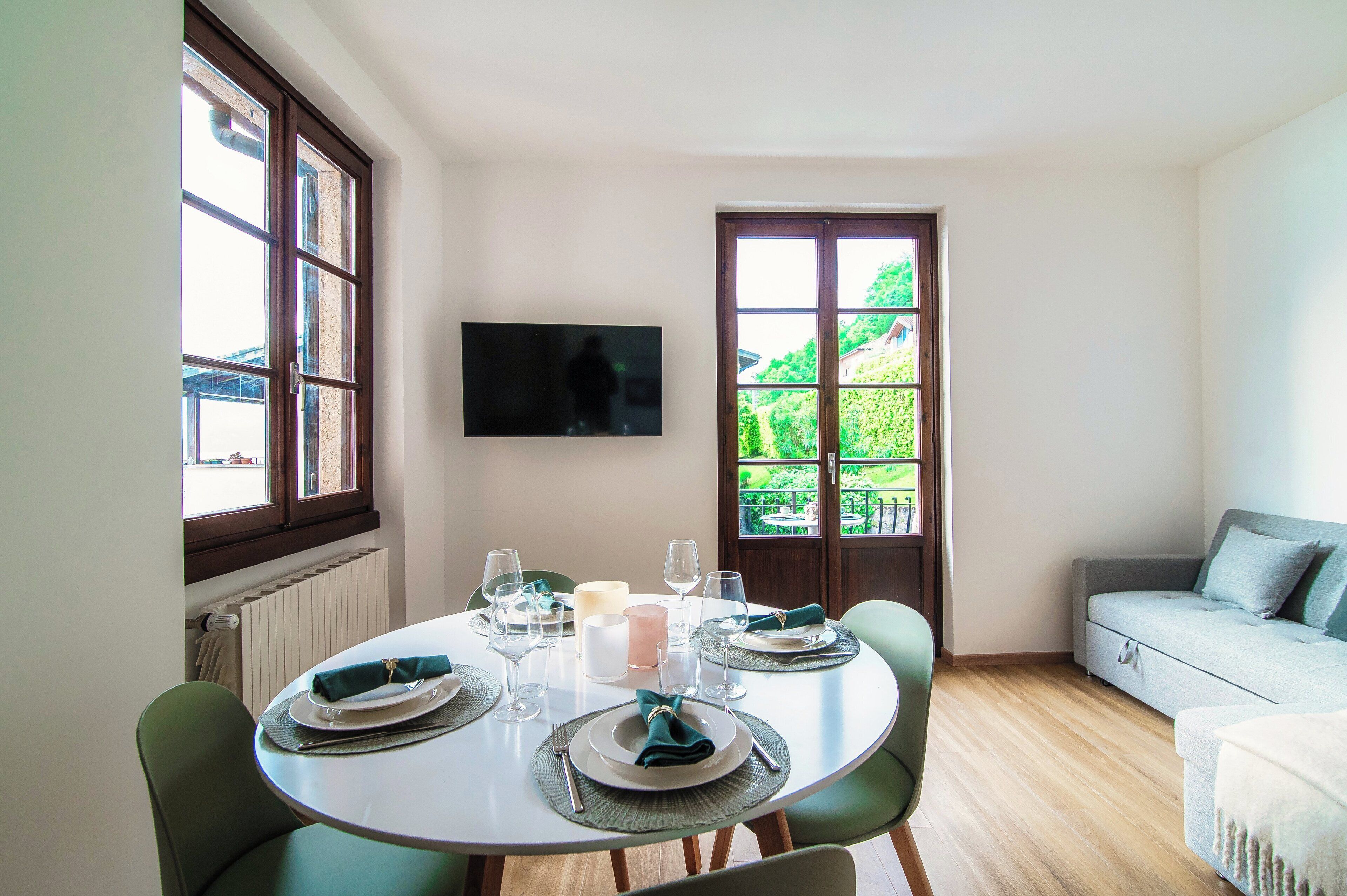 Un comedor con una mesa redonda dispuesta para cuatro personas, un televisor montado en la pared y vistas a zonas verdes a través de una ventana.