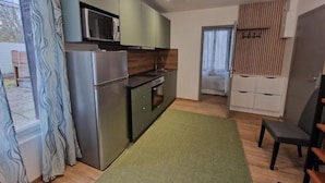 A compact kitchen with a refrigerator, microwave, and stove, a small dining area with a chair, and a view of a bedroom through an open door. - Appartement 3 (Viry-Châtillon)