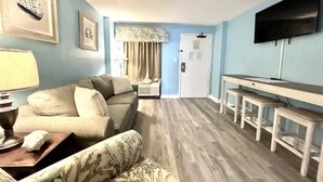 A living room with a sofa, a table, and a TV. There is a door in the background. - Amazing Oceanfront 1 Bedroom (Myrtle Beach)
