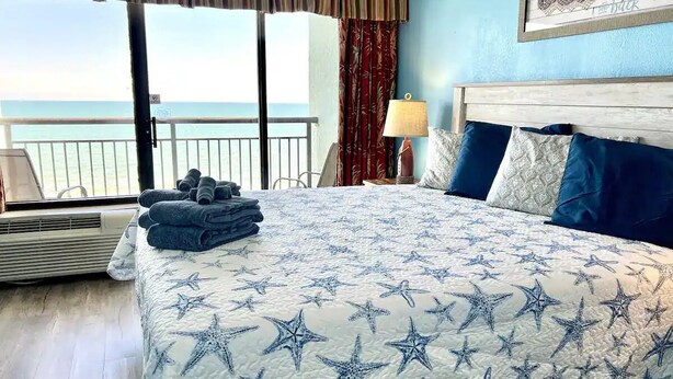 A hotel room with a large bed, a view of the sea, a balcony, and a beach-themed wall decoration. - Amazing Oceanfront 1 Bedroom (Myrtle Beach)