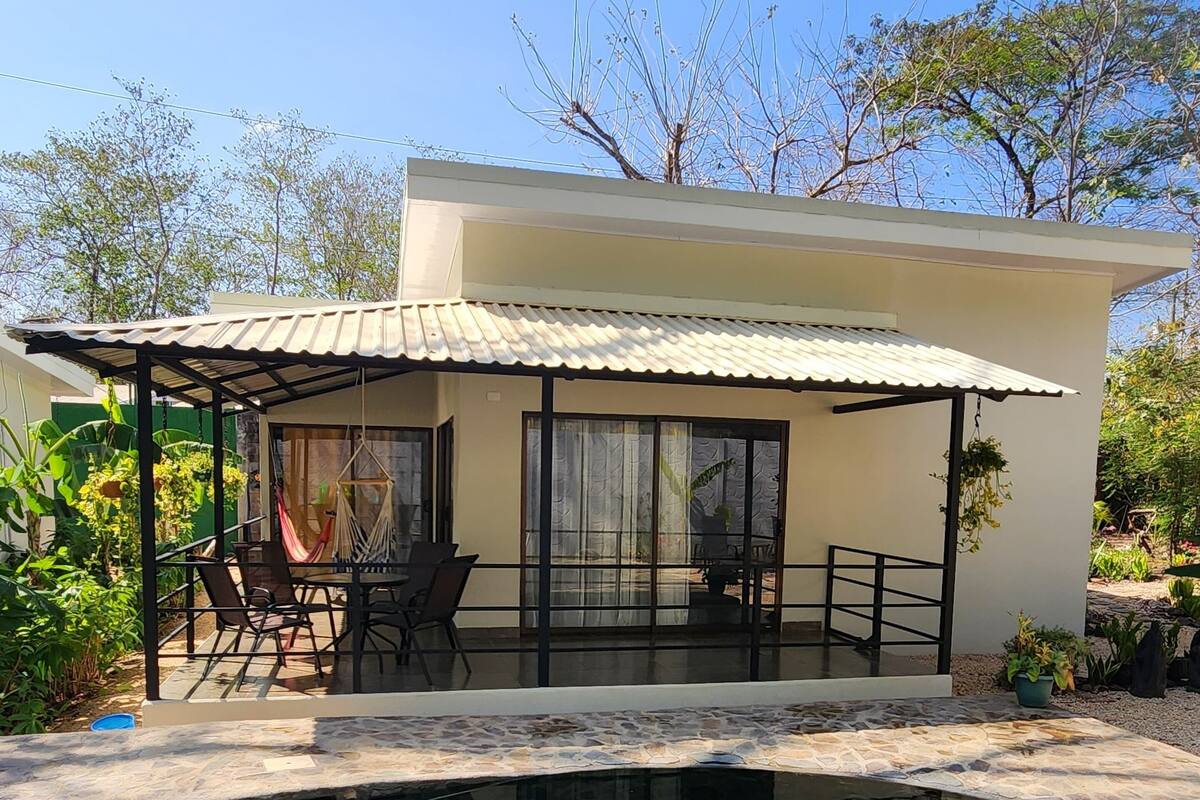Una casa moderna con piscina, un porche cubierto con mobiliario exterior y abundante vegetación alrededor.