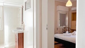 A hotel room with a wooden floor, a bed with white bedding, a bedside table, a hanging light fixture, and a bathroom with a sink and mirror. - Alimos Sea View Family Apartment (Alimos)