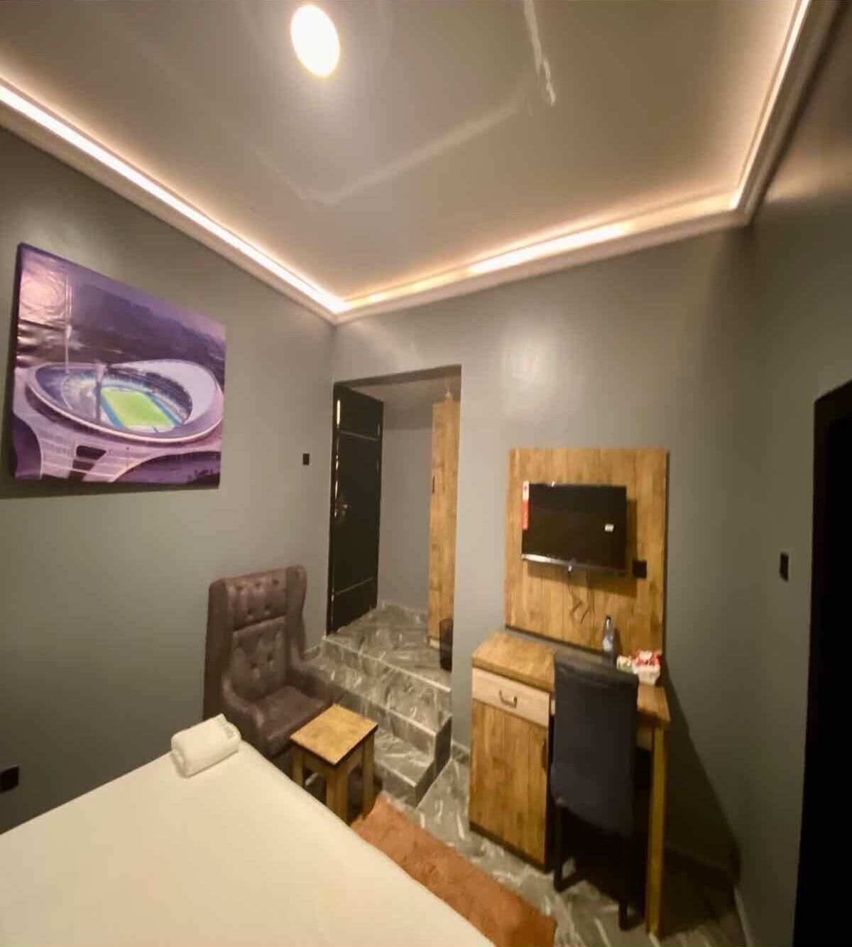 Habitación de hotel con cama, escritorio con silla, televisor montado en un panel de madera y un cuadro de un estadio en la pared.