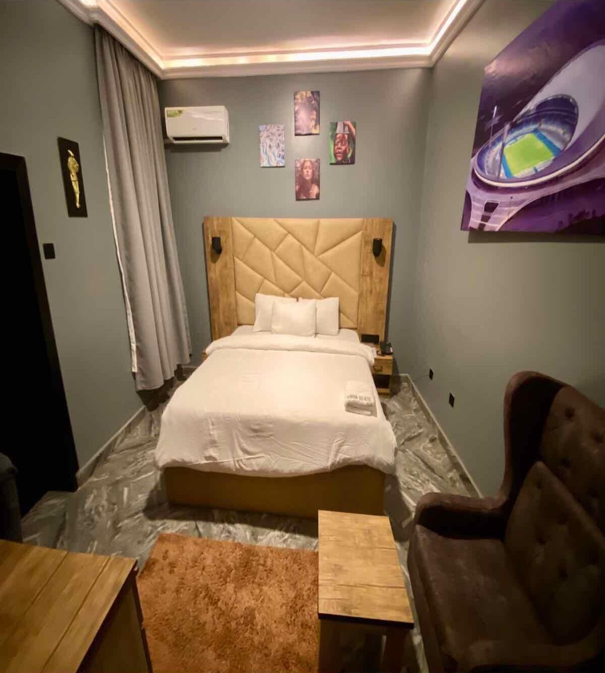 Una habitación de hotel con una cama grande, cabecera de madera, una mesita pequeña de madera, un sillón marrón y un aire acondicionado instalado en la pared.