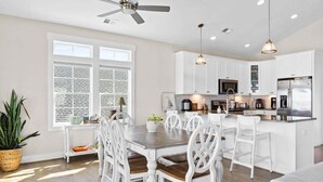 Une cuisine moderne avec des meubles blancs, un îlot central doté de sièges, un ventilateur de plafond, des luminaires suspendus et une grande fenêtre avec des stores.