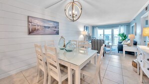 A dining area with a glass table, white chairs, and a chandelier. There is a painting on the wall and a view of the outdoors through large windows. - Intracoastal and Gulf of Mexico water views from this hideaway unit complete with heated pool, hot tub & home office (Indian Shores)