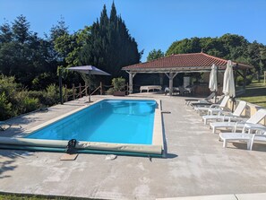 A rectangular swimming pool with a covered patio area, lounge chairs, and a barbecue setup.