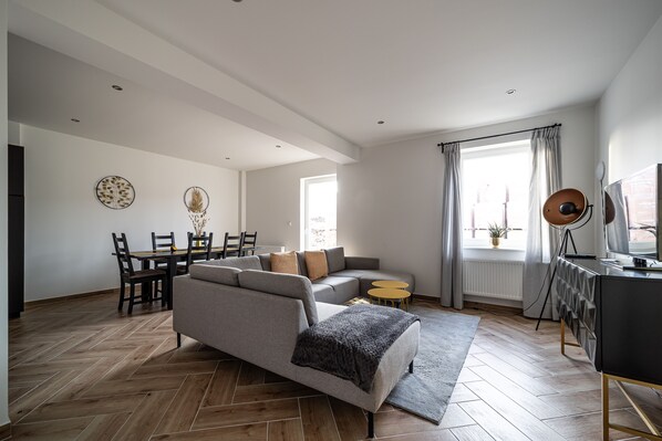 Ein modernes Wohnzimmer mit einer Couch, einem Esstisch und Stühlen. Es gibt ein großes Fenster mit Vorhängen, einen kleinen runden Tisch und ein TV-Möbel.