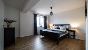 A bedroom with a bed, bedside lamps, a window with curtains, a wooden floor, and a ceiling light fixture.