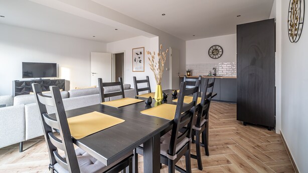 A modern living room with a sofa, a dining table with chairs, and a kitchen area with a refrigerator and a clock on the wall.