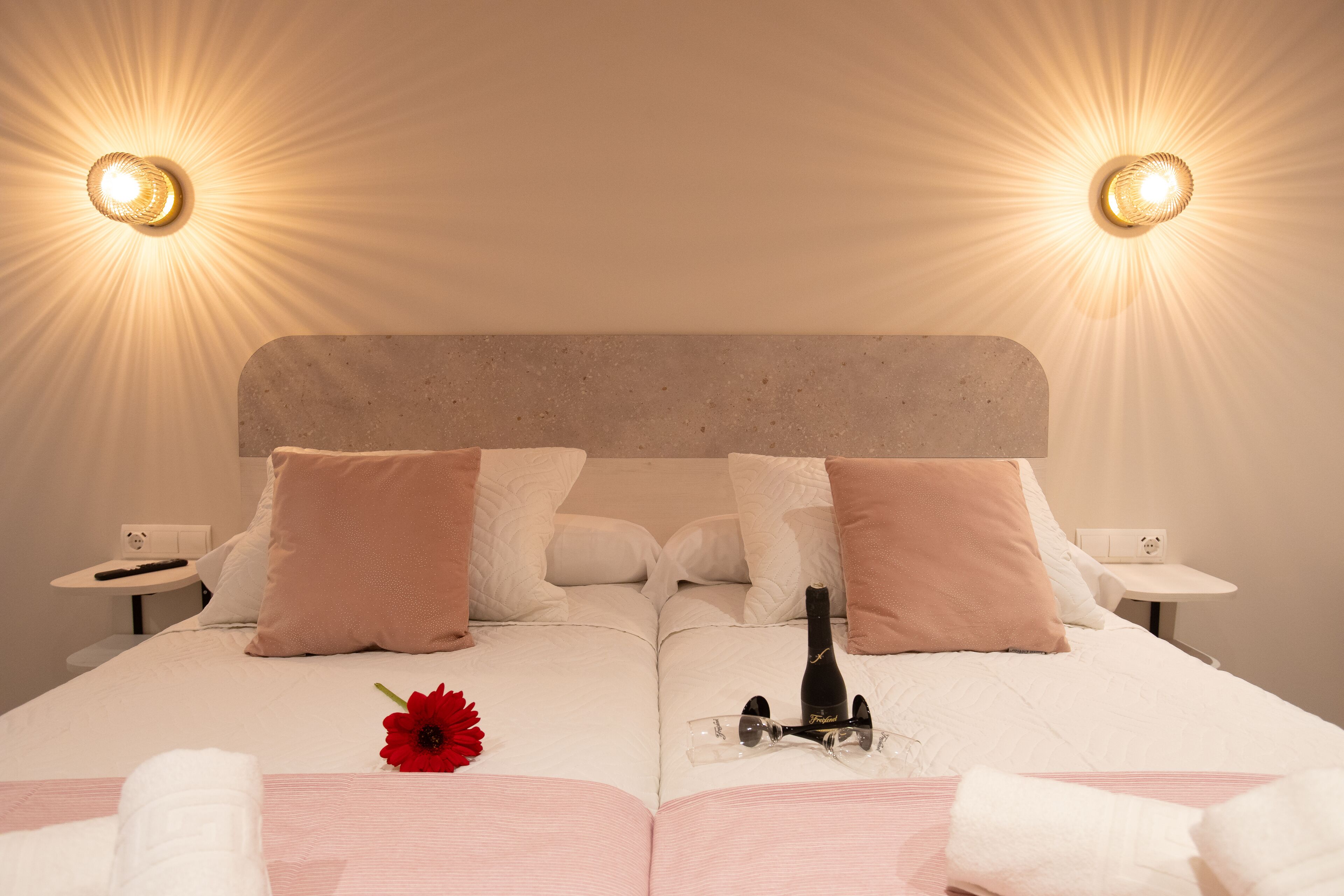 Uma cama de casal com roupa de cama branca e rosa, uma garrafa de champanhe e uma flor vermelha ao lado esquerdo.