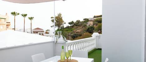 A white balcony with a table and chairs, a bottle, and two glasses on a mat.