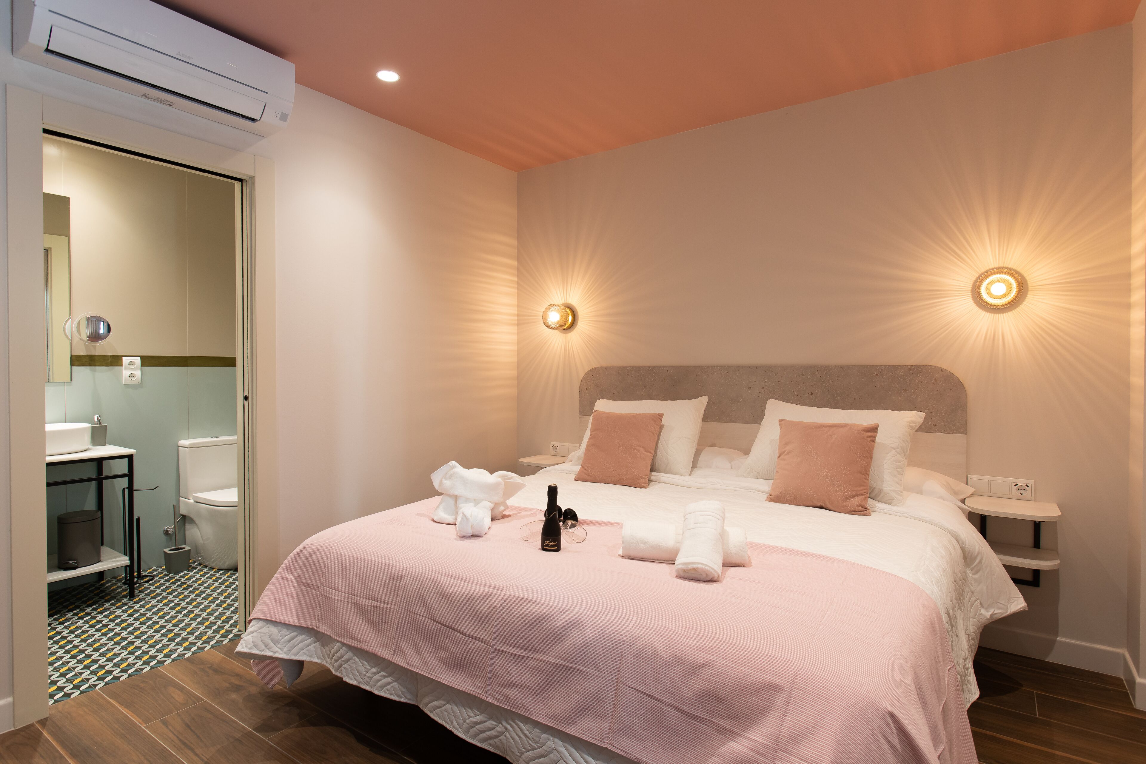A hotel room with a bed, bedside tables, a nightstand, a wall-mounted light, and a bathroom visible through an open door.