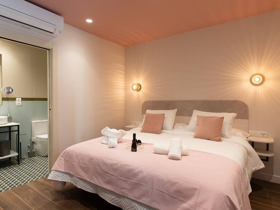 A hotel room with a bed, bedside tables, a nightstand, a wall-mounted light, and a bathroom visible through an open door.