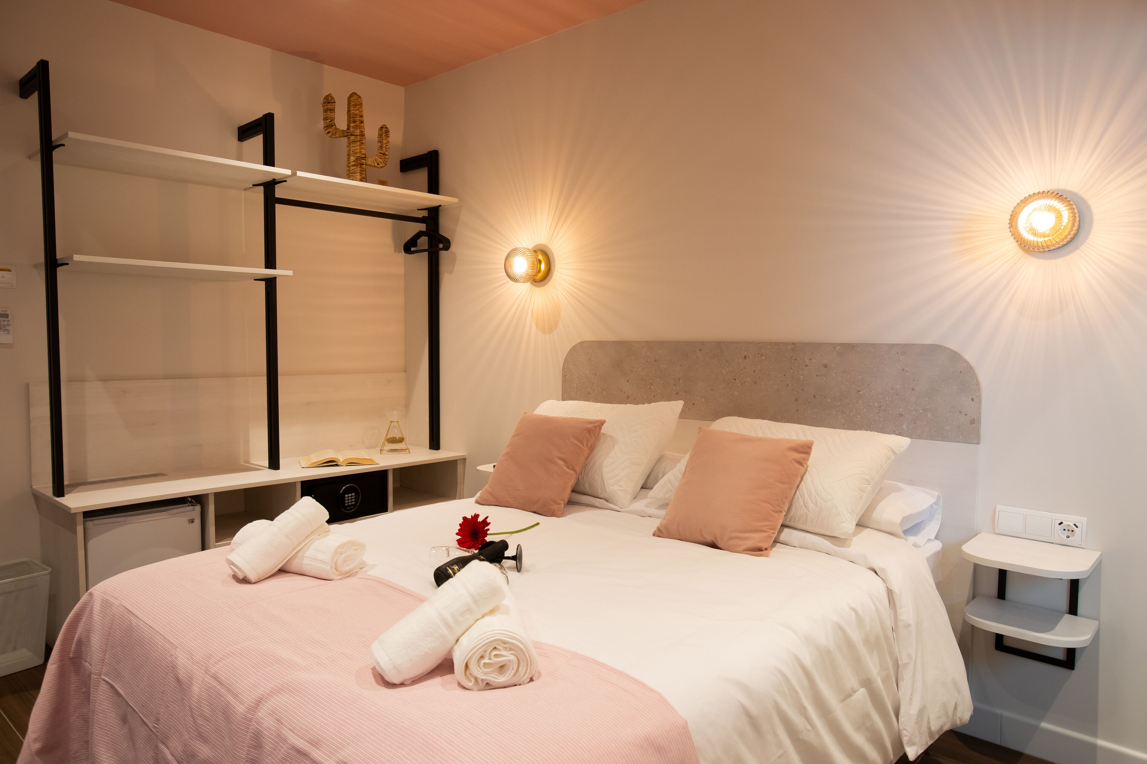 Una habitación de hotel ordenada con cama, mesitas de noche, tocador con espejo e iluminación en la pared.