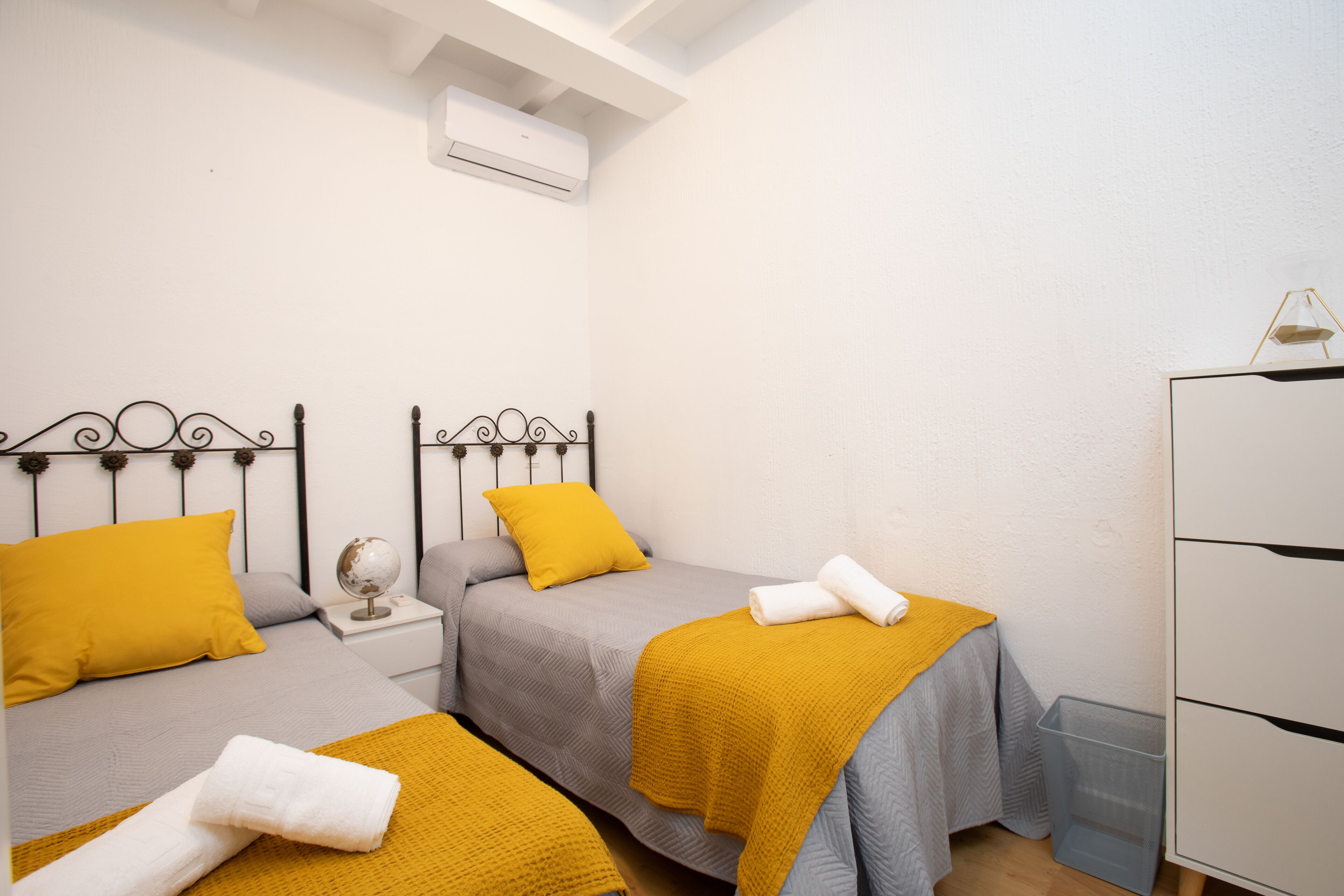 Un dormitorio con dos camas, cada una con una almohada amarilla y una toalla blanca. Hay una mesita de noche pequeña con un globo terráqueo y una lámpara.