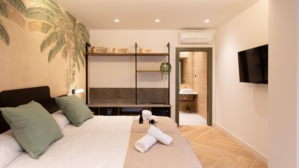 A modern hotel room with a bed, a TV, a shelf with decorative items, and a bathroom visible through an open door.