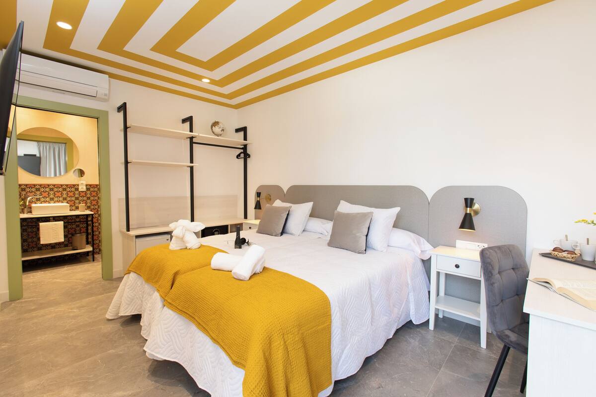A modern bedroom with a bed, bedside table, and a desk with a chair. There is a mirror, a shelf, and a small plant on the desk.