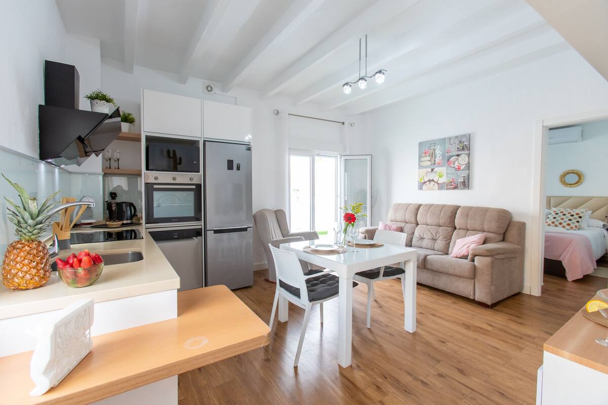 A modern kitchen with white cabinetry, a built-in oven, and a refrigerator. There is a dining area with a white table and chairs, and a living room with a beige sofa.