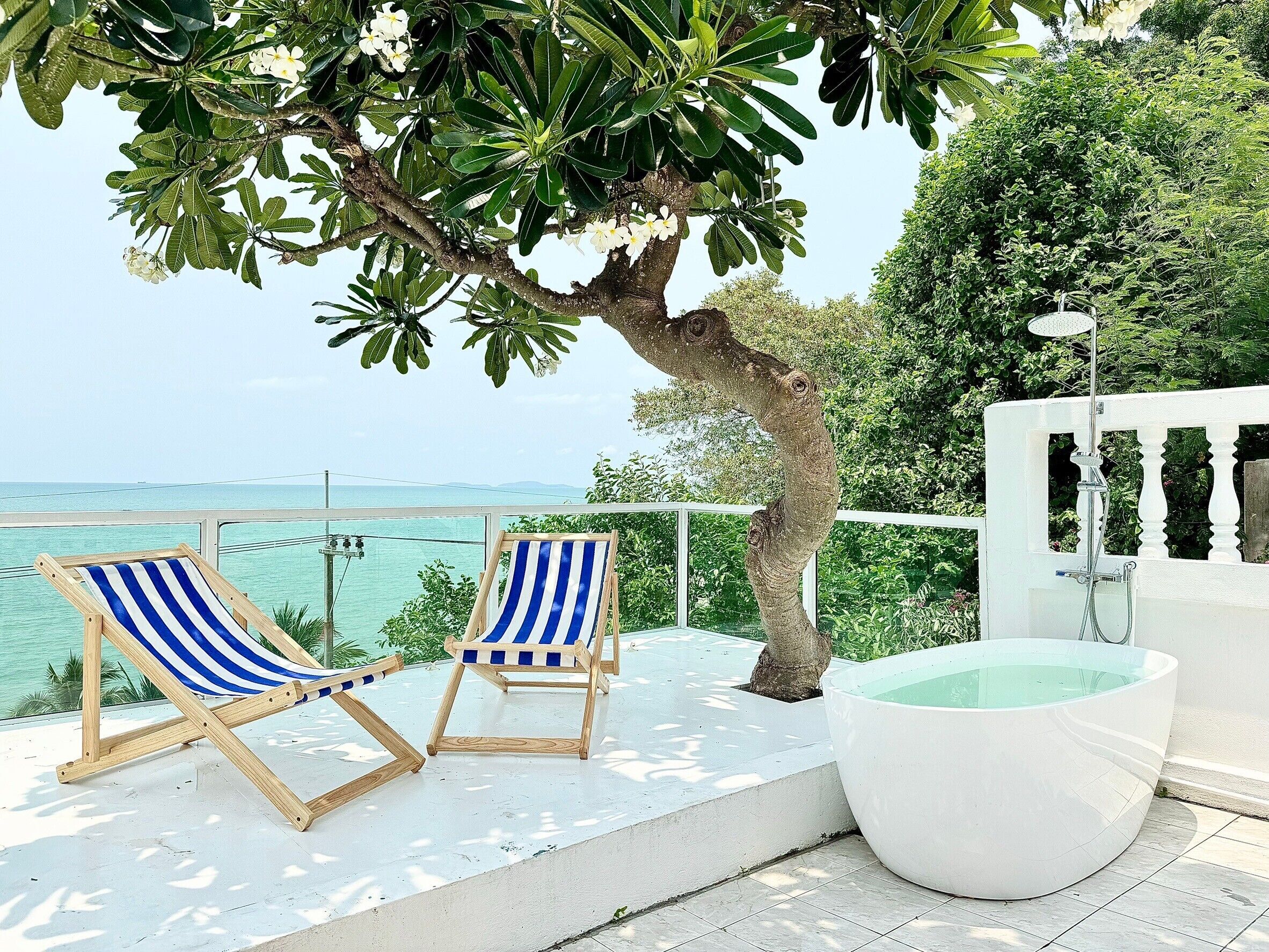 A balcony with two striped lounge chairs, a large white bathtub, and a view of the ocean.