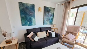 A modern living room with a black sofa, a small side table, a lamp, and a chair with a cushion. There are abstract paintings on the wall and a window with curtains. - Apartamento Junto al mar en Almuñécar, Recien Renovado y Equipado Para 4 pax (Almuñecar)