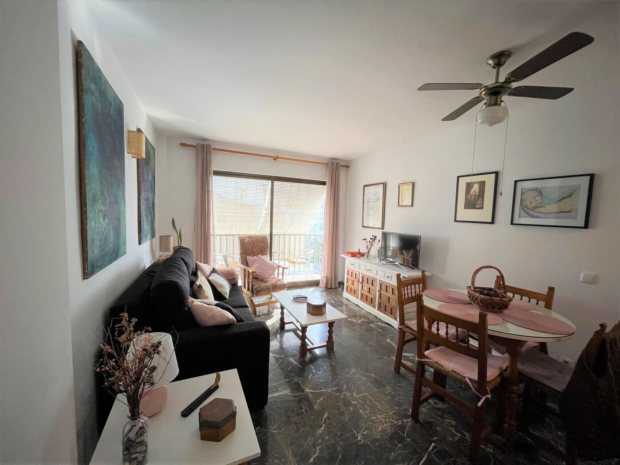 A living room with a sofa, coffee table, dining table, and chairs. There is a ceiling fan, a TV, and artwork on the walls.