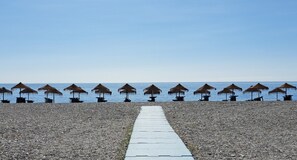 A pebble beach with rows of thatched umbrellas and tables, leading to the sea.
