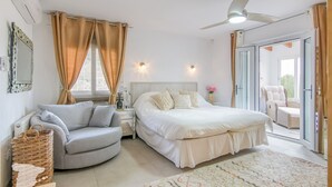 Ein Schlafzimmer mit Bett, Stuhl, Nachttisch, Fenster mit Vorhängen und Blick ins Freie.
