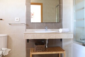 A bathroom with a white sink, a mirror above it, a wooden stool, and a shower area.