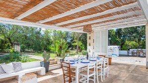 Une terrasse extérieure avec une table blanche dressée pour le repas, entourée de chaises en bois et d’un banc blanc agrémenté de coussins.