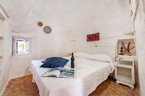 Une chambre à coucher avec un lit, une fenêtre, une table de chevet avec une bouteille et des verres, et un objet décoratif rouge accroché au mur.