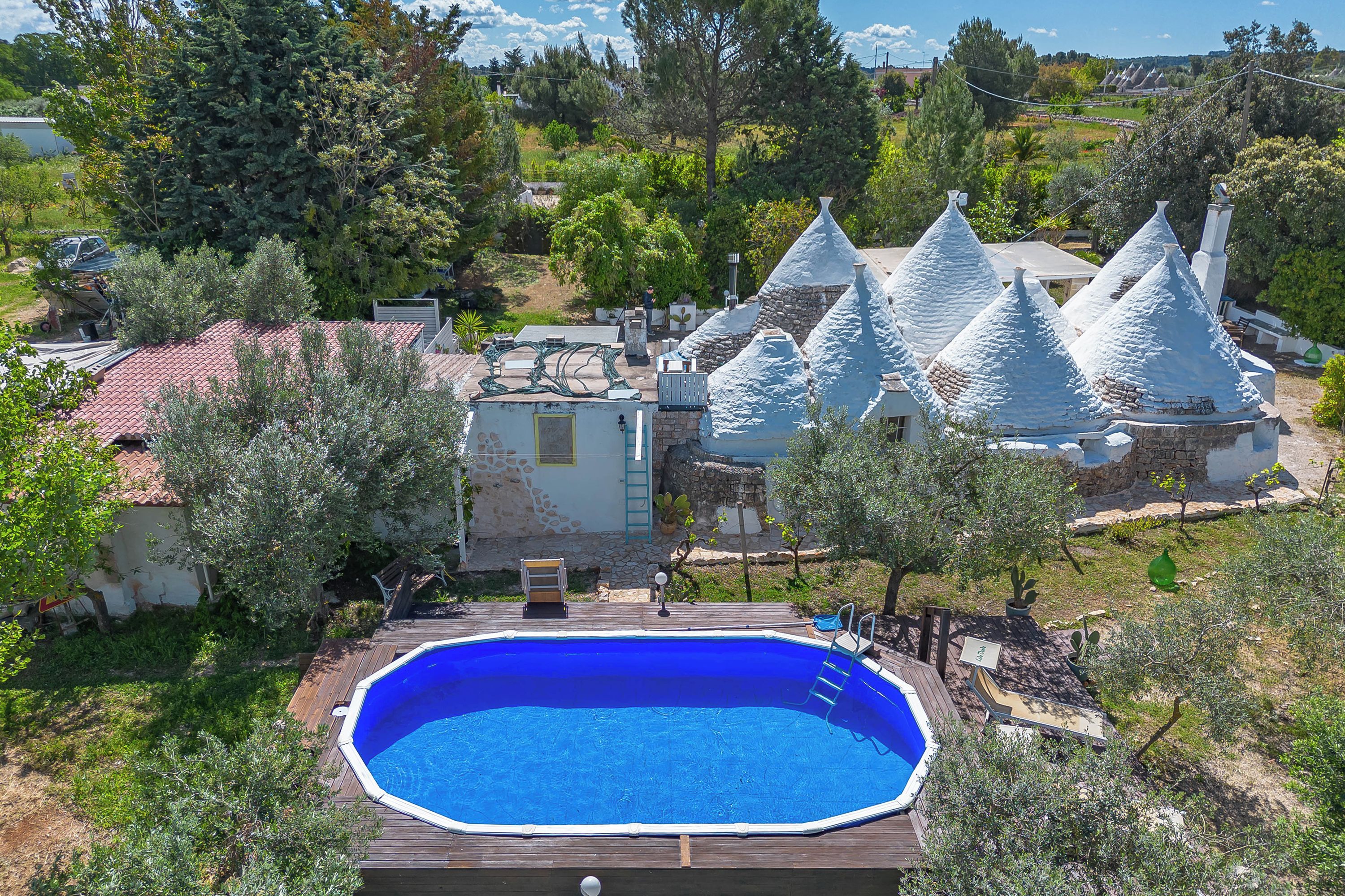 A swimming pool surrounded by a wooden deck, with a small house and conical-roofed structures nearby.