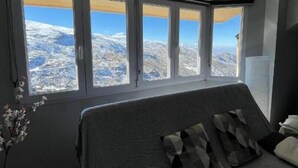 Une chambre avec un lit, une vue sur des montagnes enneigées par de grandes fenêtres et un vase rempli de fleurs.