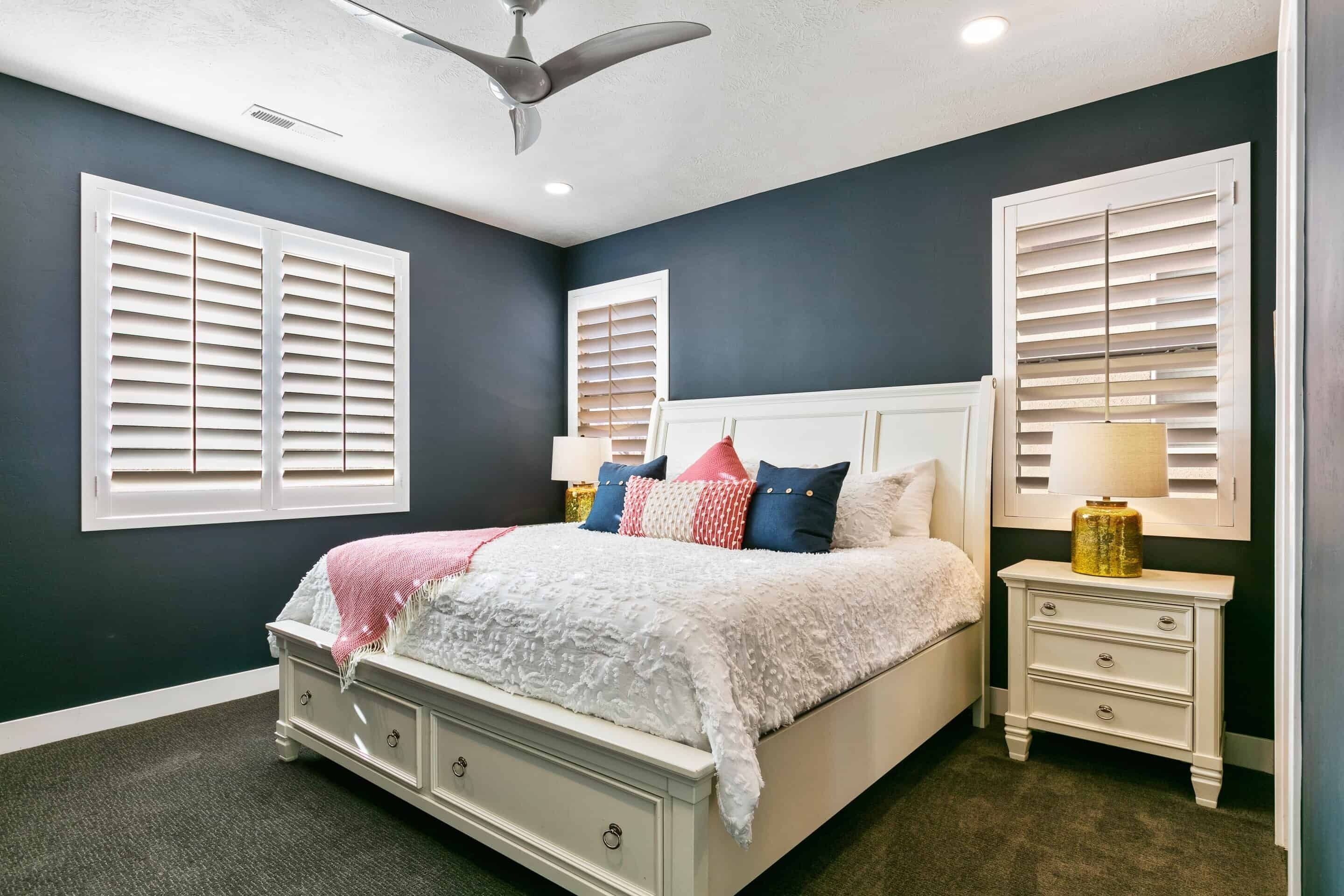 A bedroom with a bed, two nightstands, a ceiling fan, and two windows with blinds.