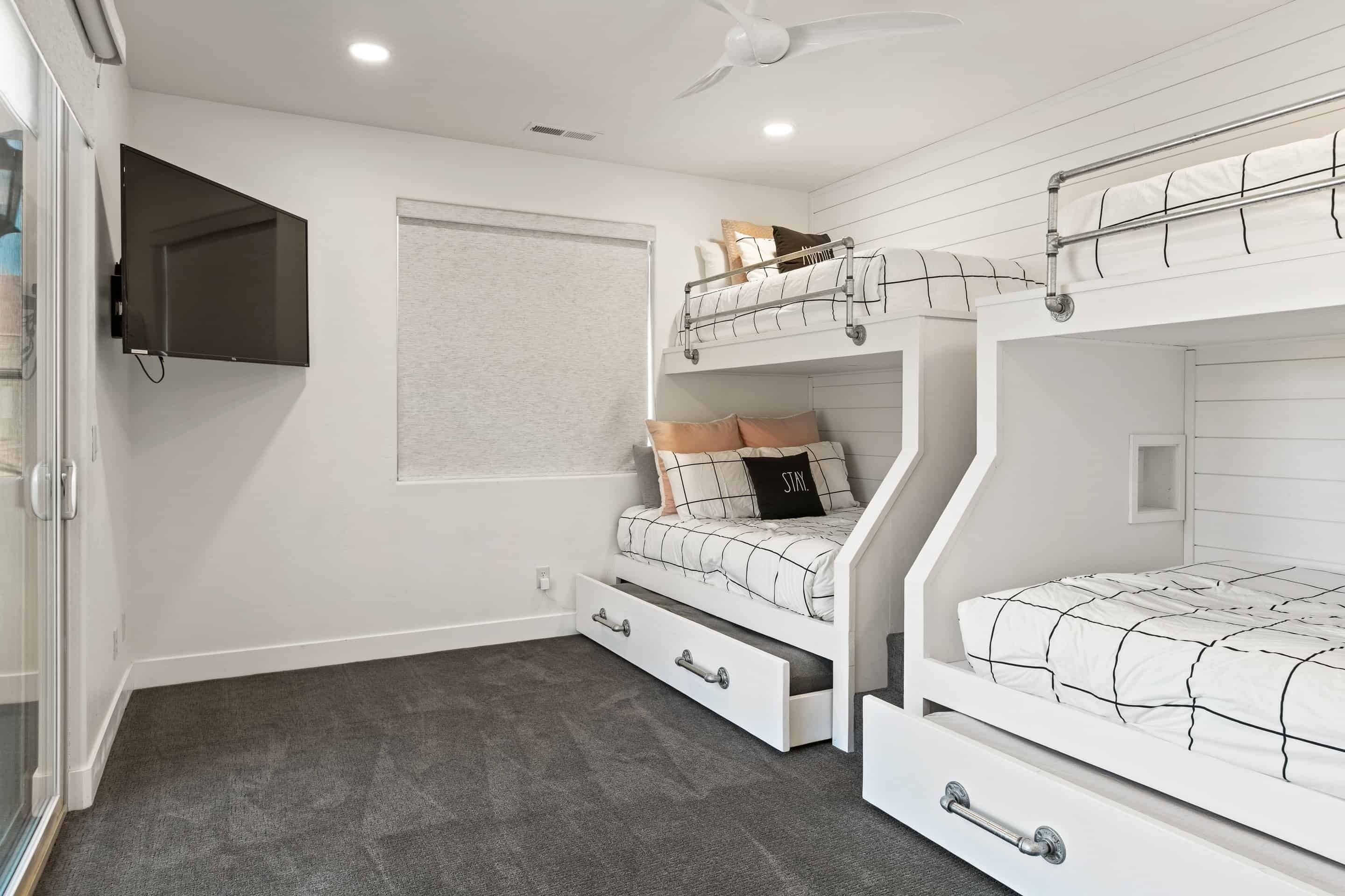 A bunk bed room with a TV mounted on the wall, a ceiling fan, and a window with blinds.