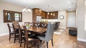 A spacious dining area with a large wooden table and multiple chairs, a kitchen with wooden cabinets, and a modern chandelier. - Double The Fun 7 Bedroom Home (Ivins)