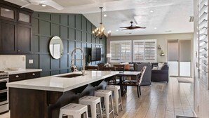 A modern kitchen with dark cabinetry, a large island with stools, a ceiling fan, and a dining area with a sofa and chairs. - Double The Fun 7 Bedroom Home (Ivins)