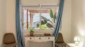 Una habitación con cama, dos sillas, un escritorio con cajones, ventana con cortinas, lámpara y planta.