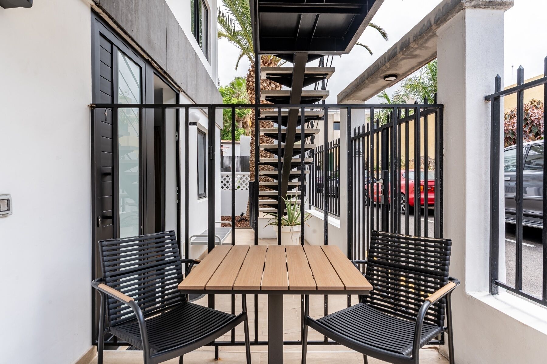 Un patio con una mesa de madera y dos sillas negras, rodeado por una cerca de metal.