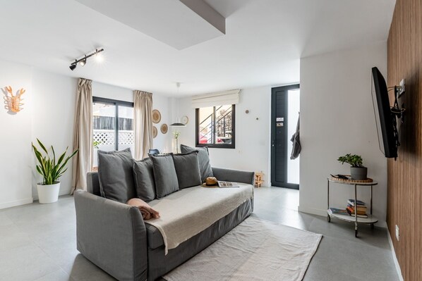 Una sala moderna con un sofá gris, una mesita auxiliar con libros, una planta en maceta y vistas al exterior a través de una ventana.