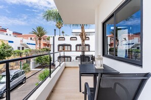 Un balcón con mesa y sillas, con vista a una calle con coches aparcados y edificios.