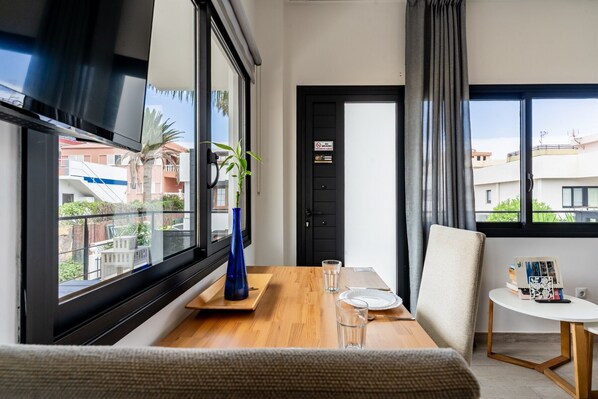 A modern hotel room with a wooden dining table, a blue vase, and a view of the cityscape through large windows.