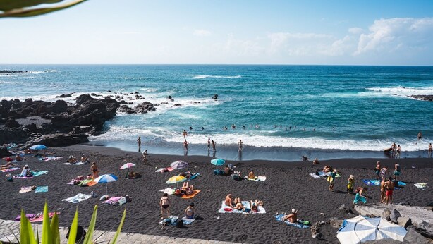 Una playa con gente tomando el sol y nadando, costa rocosa y mar azul cristalino.